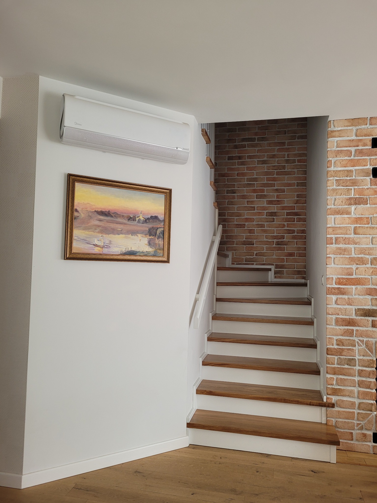 Nowoczesne schody z drewnianymi stopniami i ceglaną ścianą, oświetlone naturalnym światłem. Na ścianie klimatyzator i obraz w złotej ramie.