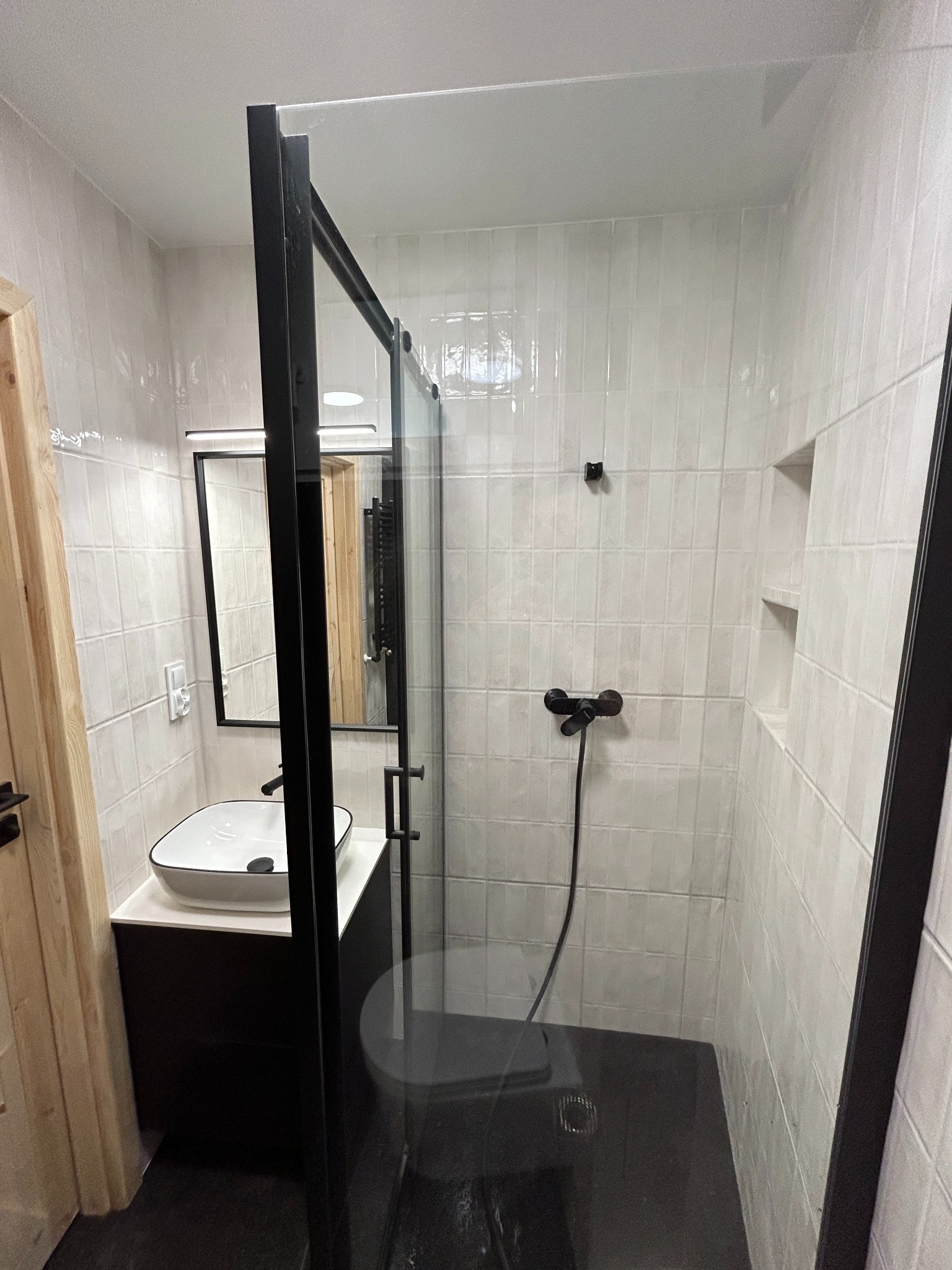 Nowoczesna łazienka z kabiną prysznicową typu walk-in z czarnymi ramami, białą umywalką nablatową i czarną armaturą na tle białych, prostokątnych płytek.