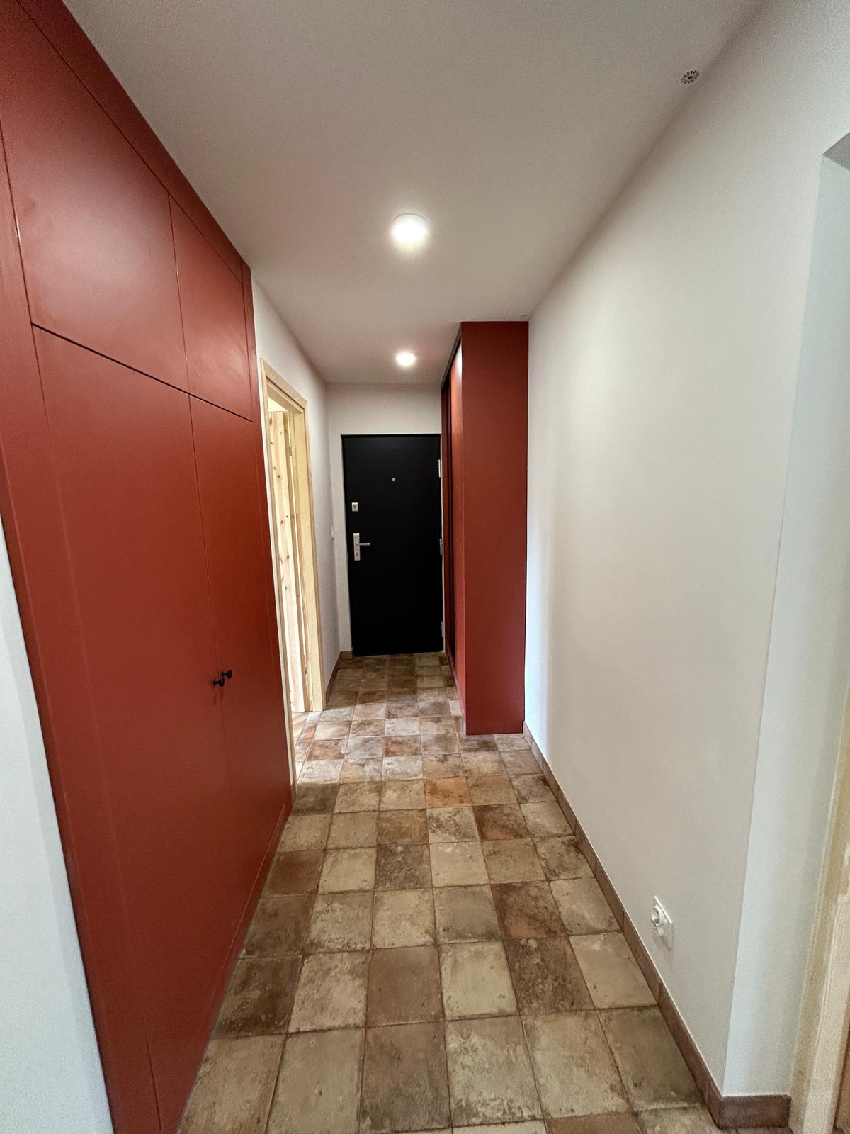 Korytarz po remoncie: terakota, zabudowa w kolorze terakoty, czarne drzwi wejściowe. Widok na wprost, oświetlenie sufitowe.