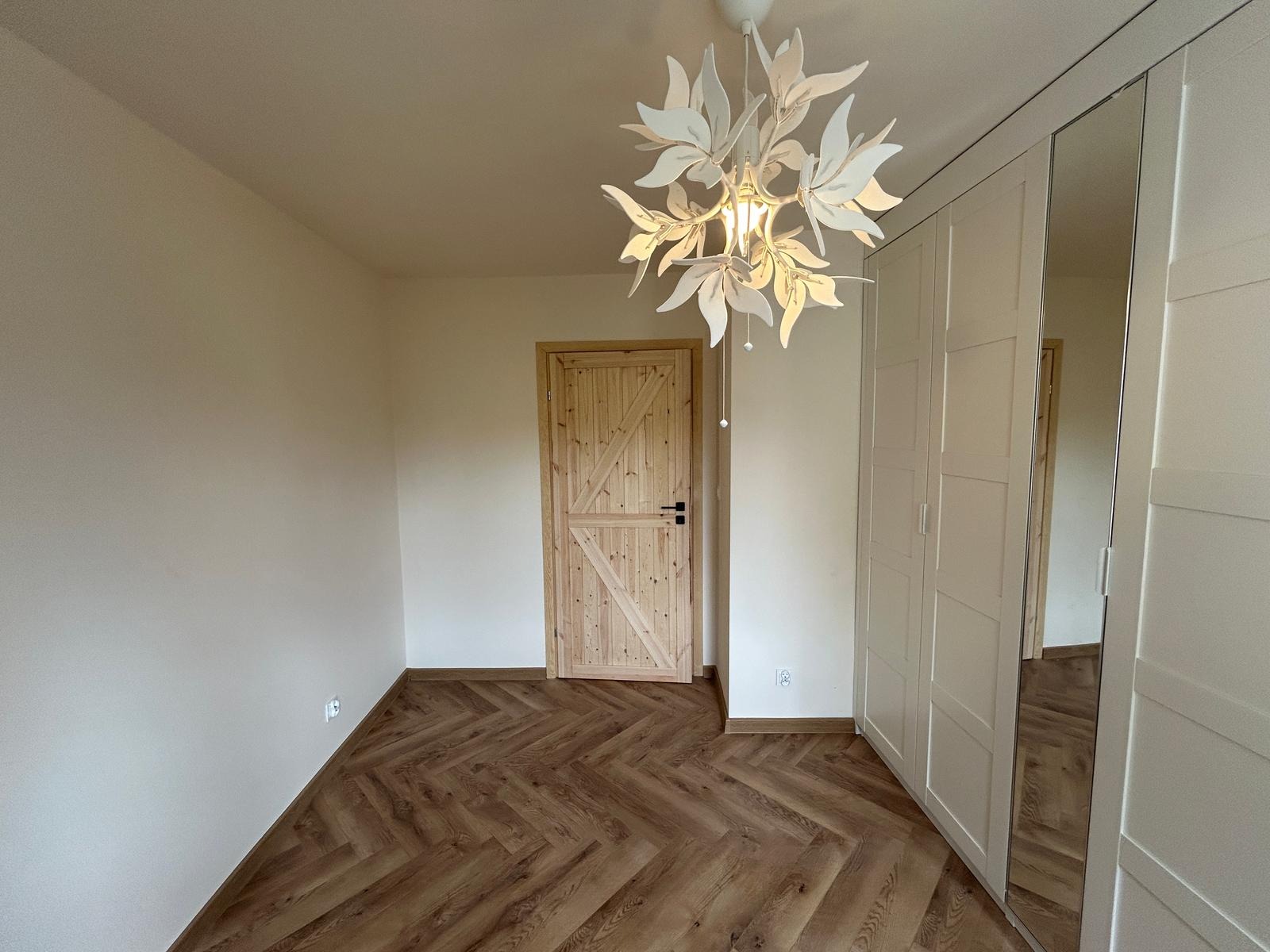 Jasne wnętrze z parkietem w jodełkę, drewnianymi drzwiami i białą szafą wnękową z lustrem. Dekoracyjna lampa sufitowa w kształcie liści.