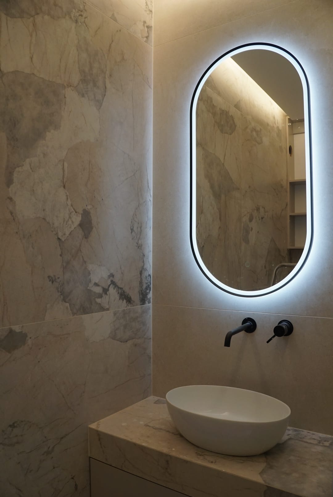 Elegancka łazienka z owalnym lustrem LED, marmurowymi płytkami i białą umywalką nablatową. Czarna armatura dodaje kontrastu. Widoczne odbicie wnętrza w lustrze.