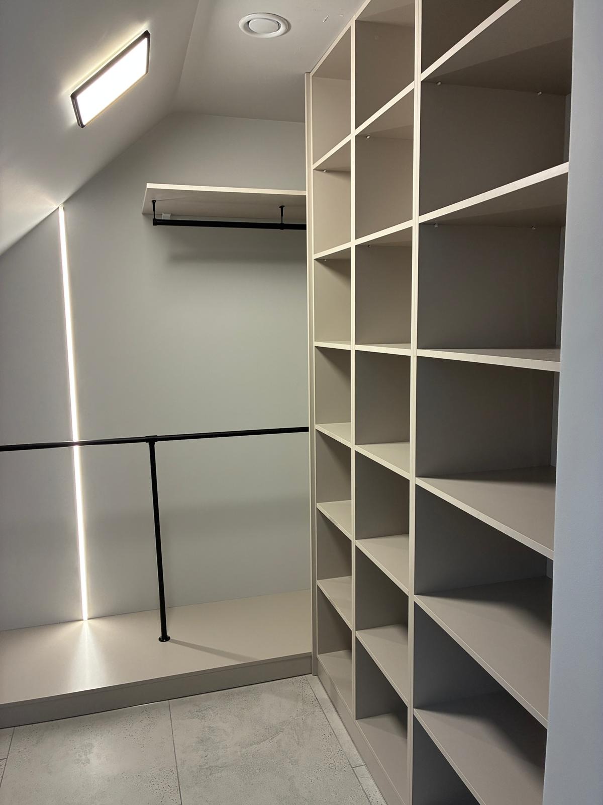 Półki wnękowe z oświetleniem LED w garderobie na poddaszu. Czarny drążek na ubrania. Minimalistyczny design, jasne kolory, nowoczesne wykończenie.