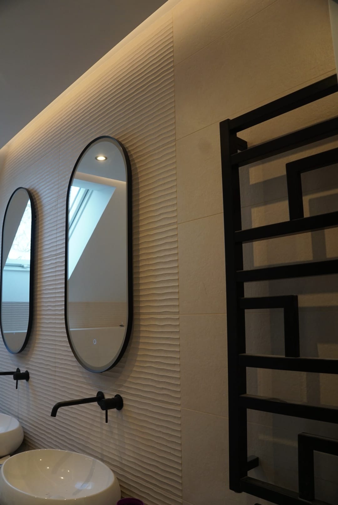 Nowoczesna łazienka z owalnymi lustrami z podświetleniem LED, czarną armaturą i grzejnikiem drabinkowym na tle ściany z dekoracyjną, falowaną fakturą.