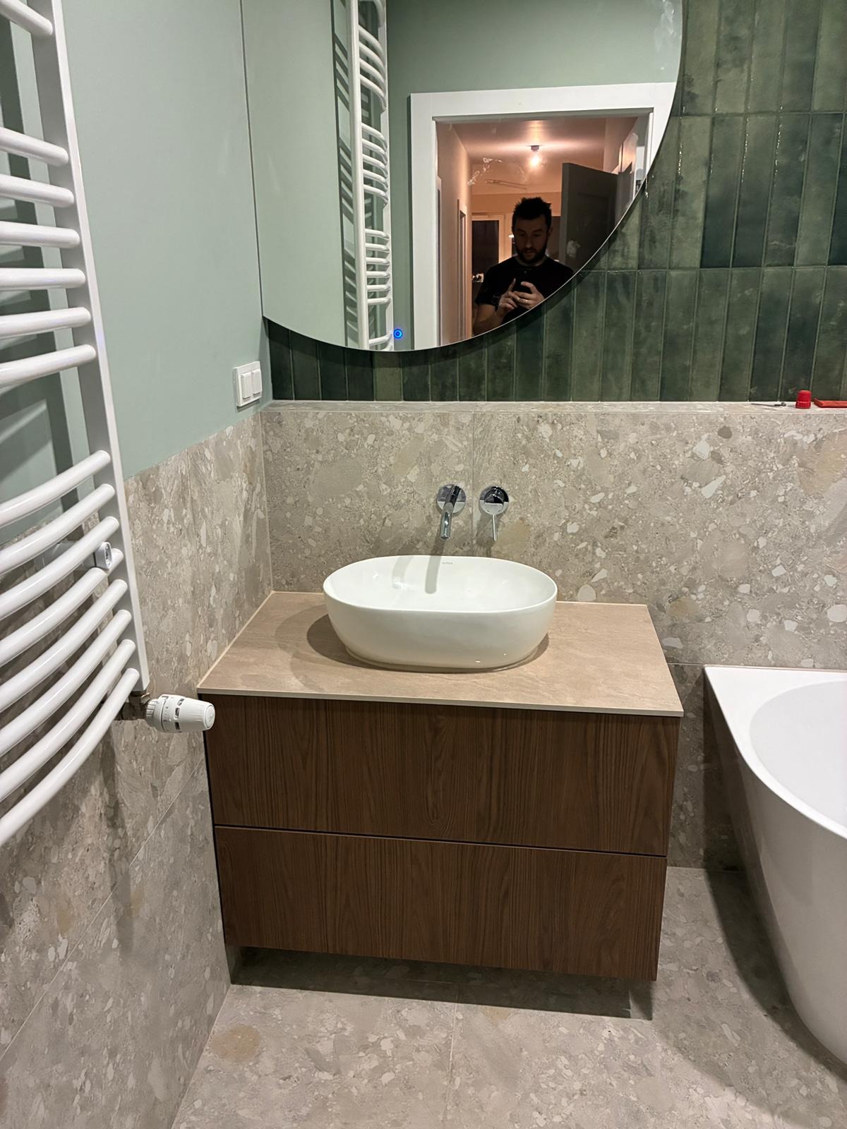 Nowoczesna łazienka z umywalką nablatową, drewnianą szafką, okrągłym lustrem z podświetleniem i grzejnikiem drabinkowym. W odbiciu lustra widoczna osoba.