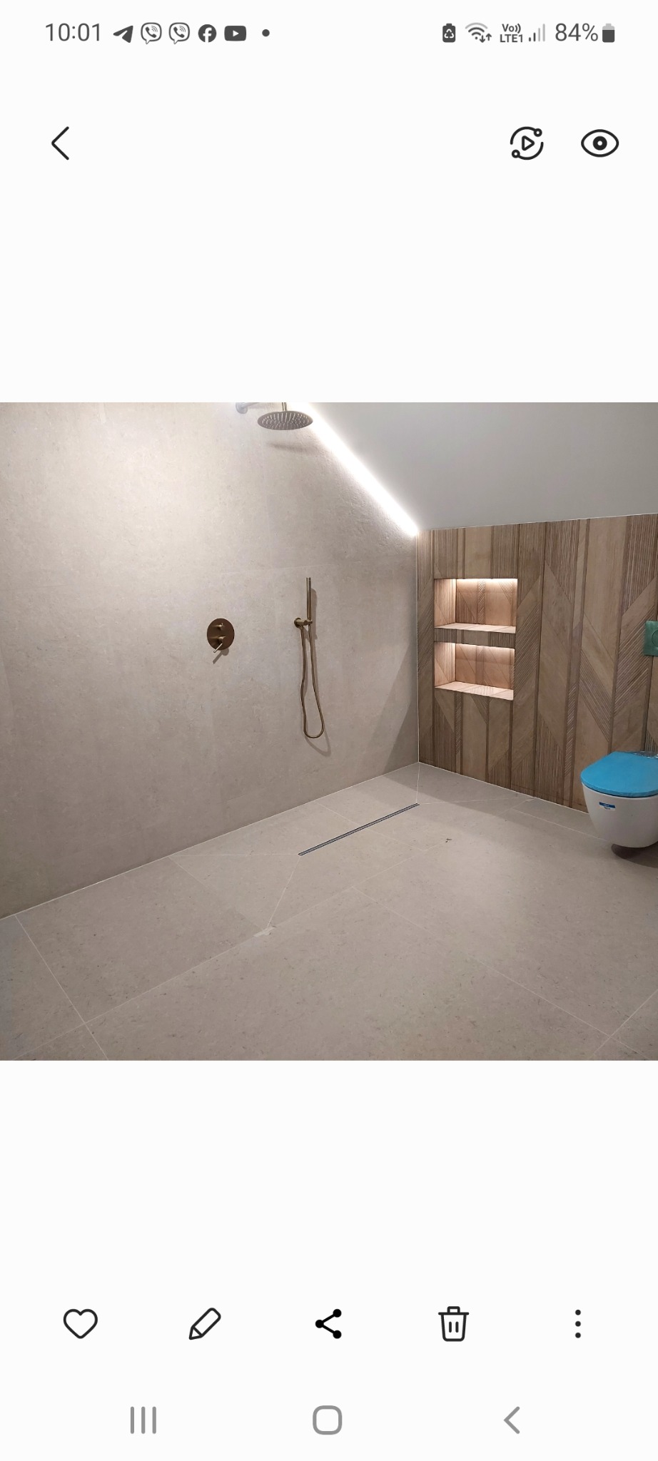 Wykończona łazienka z kabiną prysznicową typu walk-in, beżowe płytki wielkoformatowe, drewnopodobna ściana z wnękami oświetleniowymi, odpływ liniowy w podłodze i wisząca toaleta z niebieską deską.