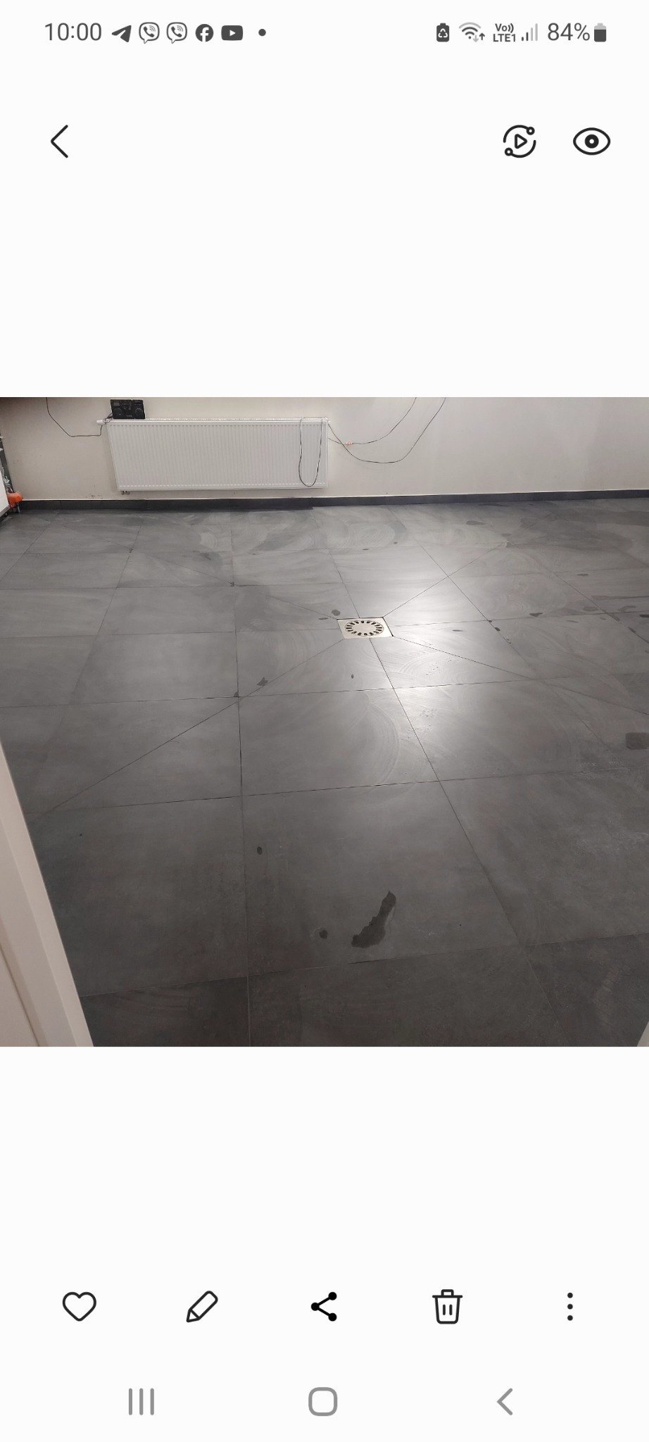 Szare, matowe płytki podłogowe ułożone w kwadraty, z odpływem liniowym pośrodku, widoczne ślady po umyciu i niedociągnięcia w czystości.