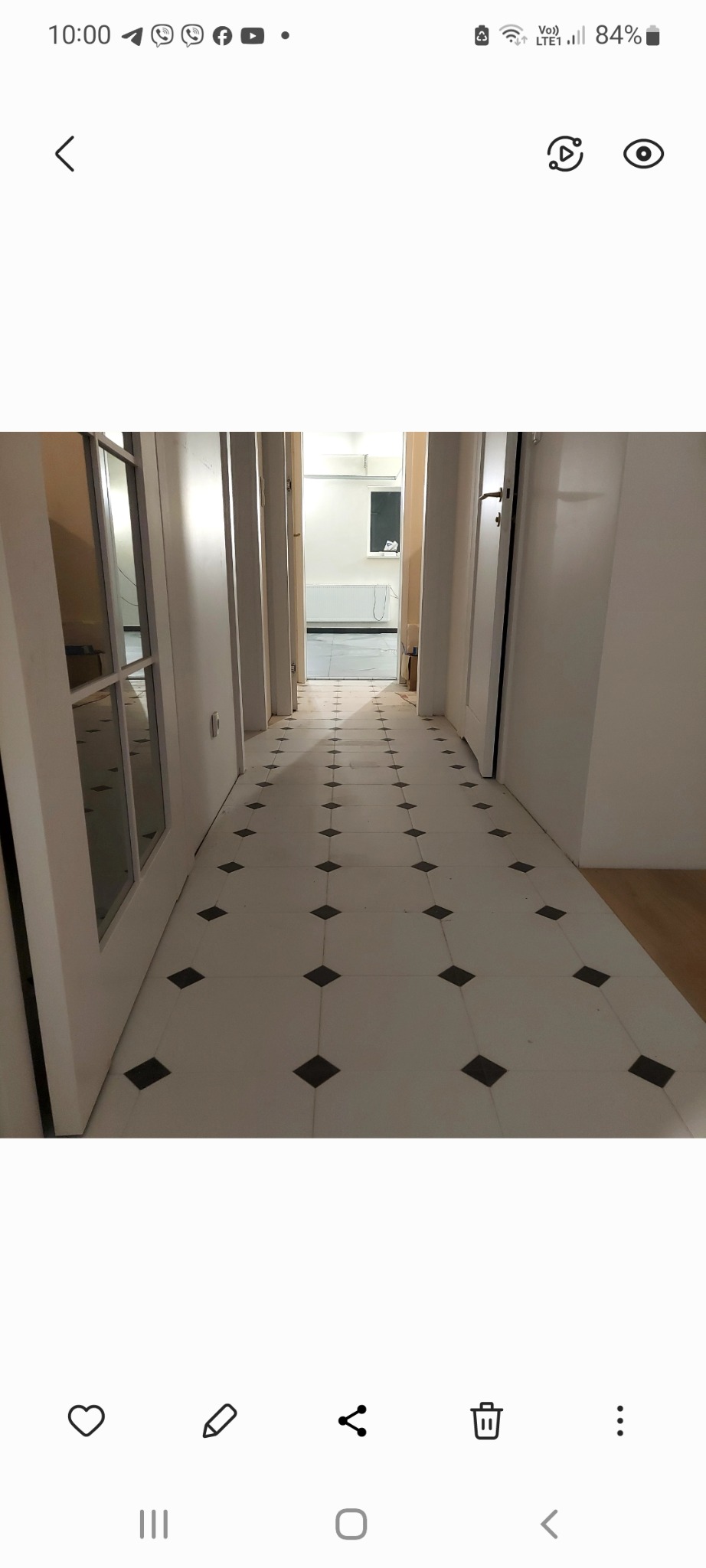 Korytarz z białymi ścianami i podłogą wyłożoną jasnymi płytkami z czarnymi rombami, widok w głąb pomieszczenia, drzwi po obu stronach.