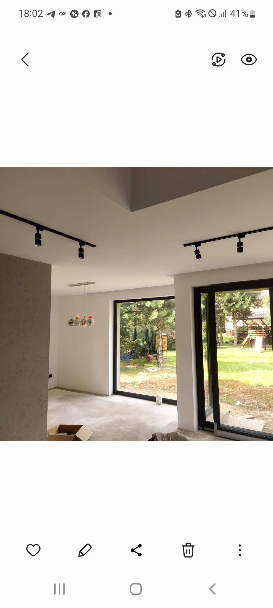 Nowoczesne oświetlenie szynowe w jasnym wnętrzu z dużymi oknami wychodzącymi na ogród. Widoczne elementy wykończenia sufitu i surowa podłoga w trakcie prac.