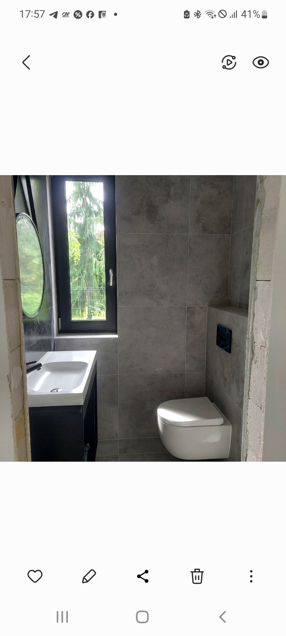 Wykończona łazienka w szarościach z wiszącą toaletą, umywalką na szafce i oknem z widokiem na zieleń. Widoczne surowe wykończenie wnęki po lewej stronie.