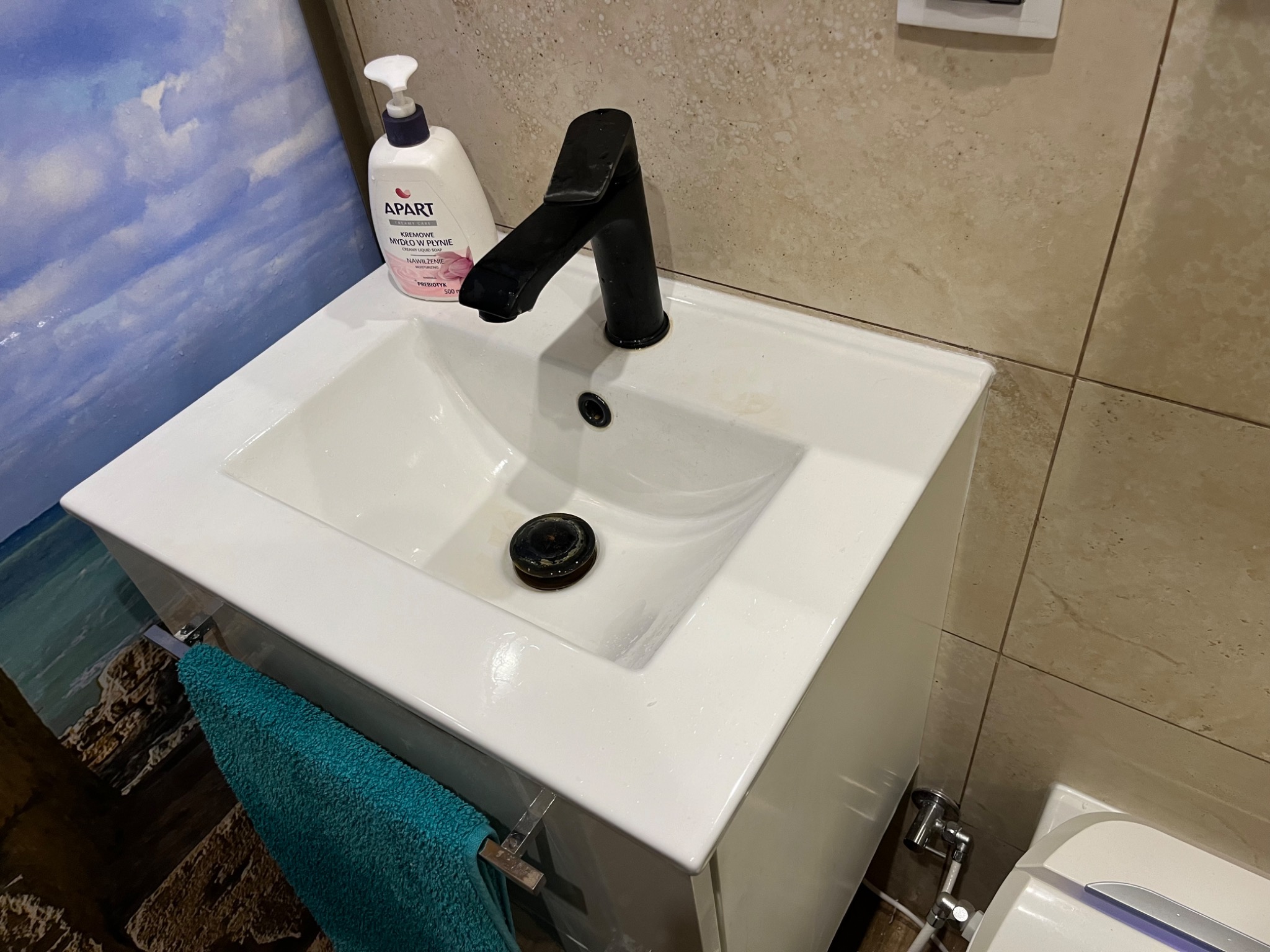 Biała umywalka z czarną baterią i dozownikiem mydła Apart na tle beżowych płytek i dekoracyjnej tapety z motywem morskim, z turkusowym ręcznikiem wiszącym z boku.