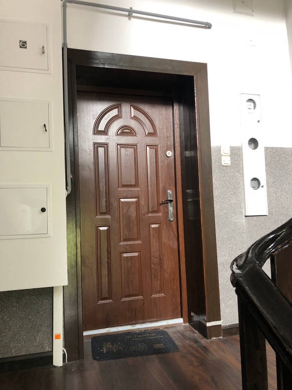 Brązowe drzwi wejściowe zamontowane w bloku, widoczne skrzynki elektryczne na ścianie i fragment klatki schodowej.