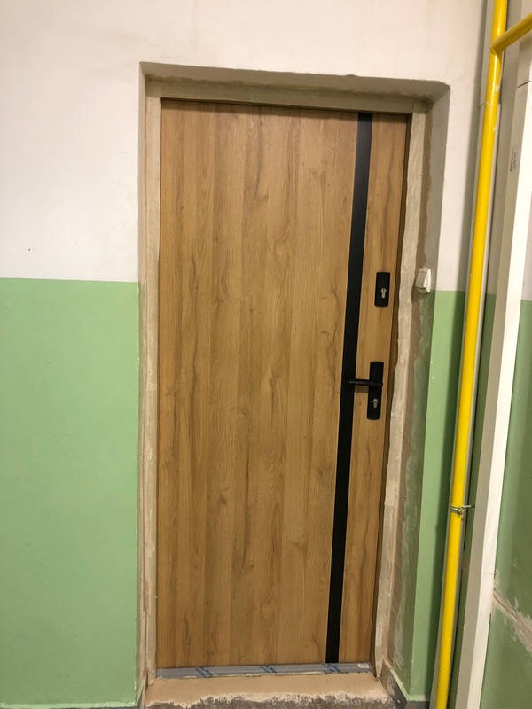 Zamontowane drzwi wejściowe z drewnopodobną okleiną i czarnym paskiem dekoracyjnym w bloku z zieloną lamperią.