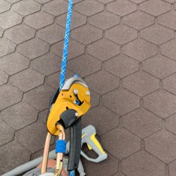 MatGras - Żółty przyrząd asekuracyjny Petzl I'D przymocowany do liny na tle brązowego dachu z gontów bitumicznych, w tle fragment myjki ciśnieniowej.