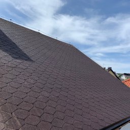 MatGras - Ciemnobrązowy dach pokryty gontem bitumicznym w kształcie rombów, widok z dołu pod kątem, fragment komina w lewym górnym rogu, błękitne niebo z chmurami w tle.