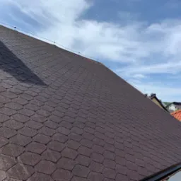 Ciemnobrązowy dach pokryty gontem bitumicznym w kształcie rombów, widok z dołu pod kątem, fragment komina w lewym górnym rogu, błękitne niebo z chmurami w tle.