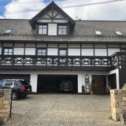 Dwukondygnacyjny dom w stylu szwajcarskim z ciemnym drewnem i białymi ścianami, dach pokryty gontem bitumicznym, podjazd z kostki granitowej, dwa samochody w garażu, numer 44 na kamiennym słupku.