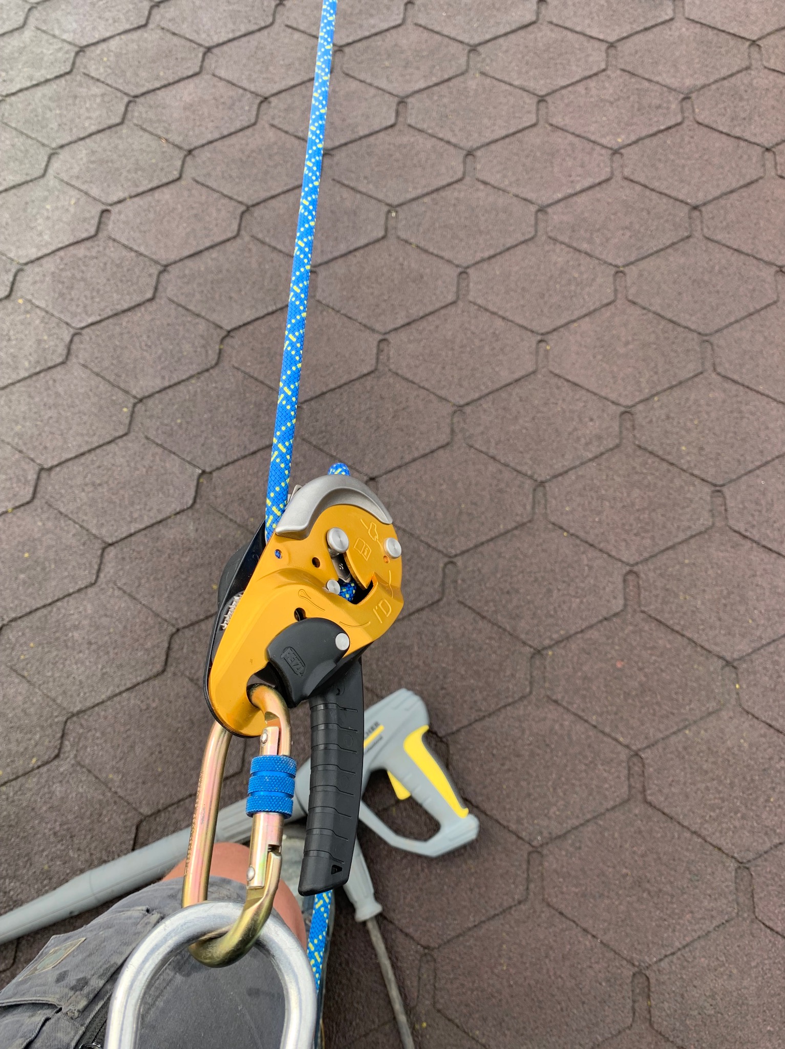 Żółty przyrząd asekuracyjny Petzl I'D przymocowany do liny na tle brązowego dachu z gontów bitumicznych, w tle fragment myjki ciśnieniowej.