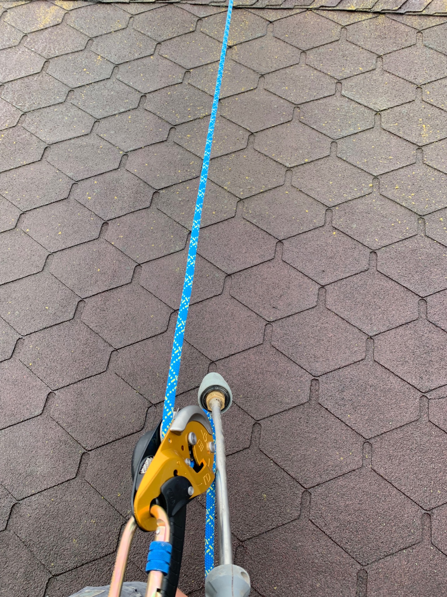 Fragment dachu pokrytego gontem bitumicznym z zamocowanym sprzętem alpinistycznym, lina asekuracyjna w kolorze niebieskim. Widoczne zabrudzenia na powierzchni dachu.