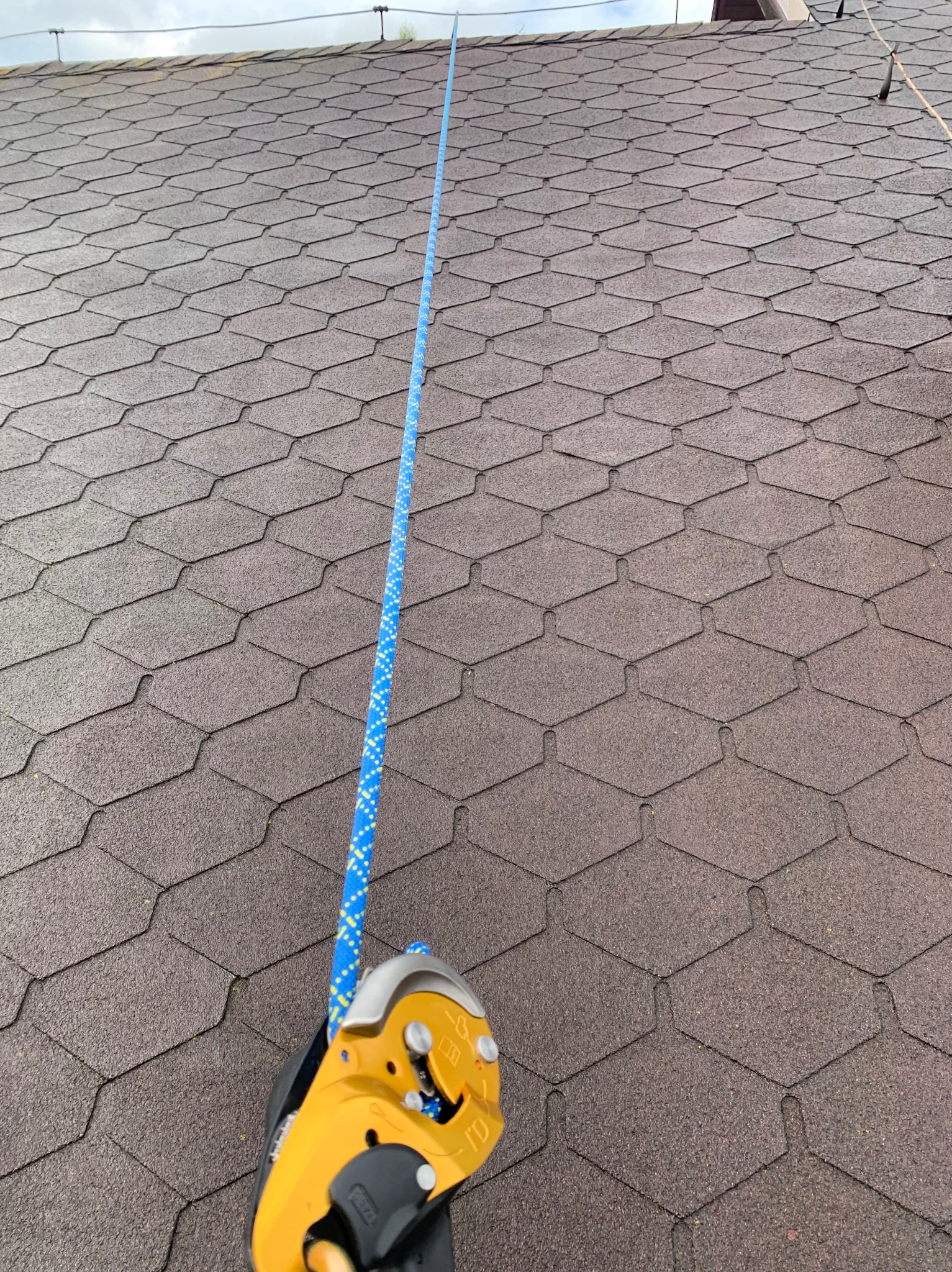 Ciemny dach pokryty gontem bitumicznym z zamocowanym, żółtym urządzeniem asekuracyjnym i niebieską liną zabezpieczającą biegnącą pionowo w górę.
