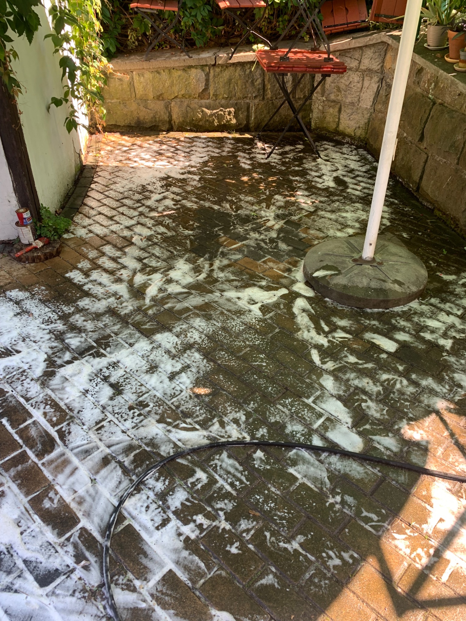 Częściowo umyta kostka brukowa na patio z kamiennym murem, widoczne resztki piany i wąż ciśnieniowy, w tle krzesła ogrodowe.