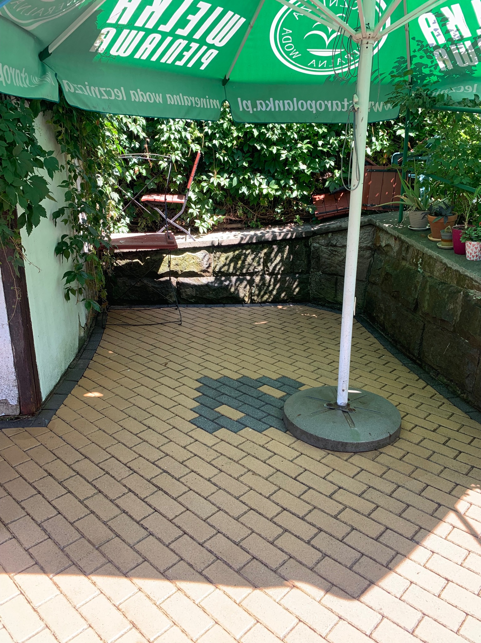 Ułożona z beżowo-żółtej kostki brukowej powierzchnia tarasu z ciemniejszym, szarym wzorem w centralnej części, ocieniona zielonym parasolem, z widocznym fragmentem ściany porośniętej bluszczem...