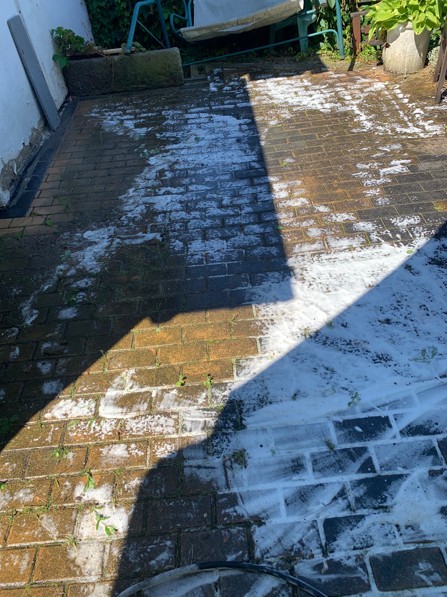 Częściowo umyta kostka brukowa z widoczną pianą czyszczącą, kontrastująca z brudną powierzchnią w cieniu, z zaroślami między kostkami i fragmentem węża na dole kadru.