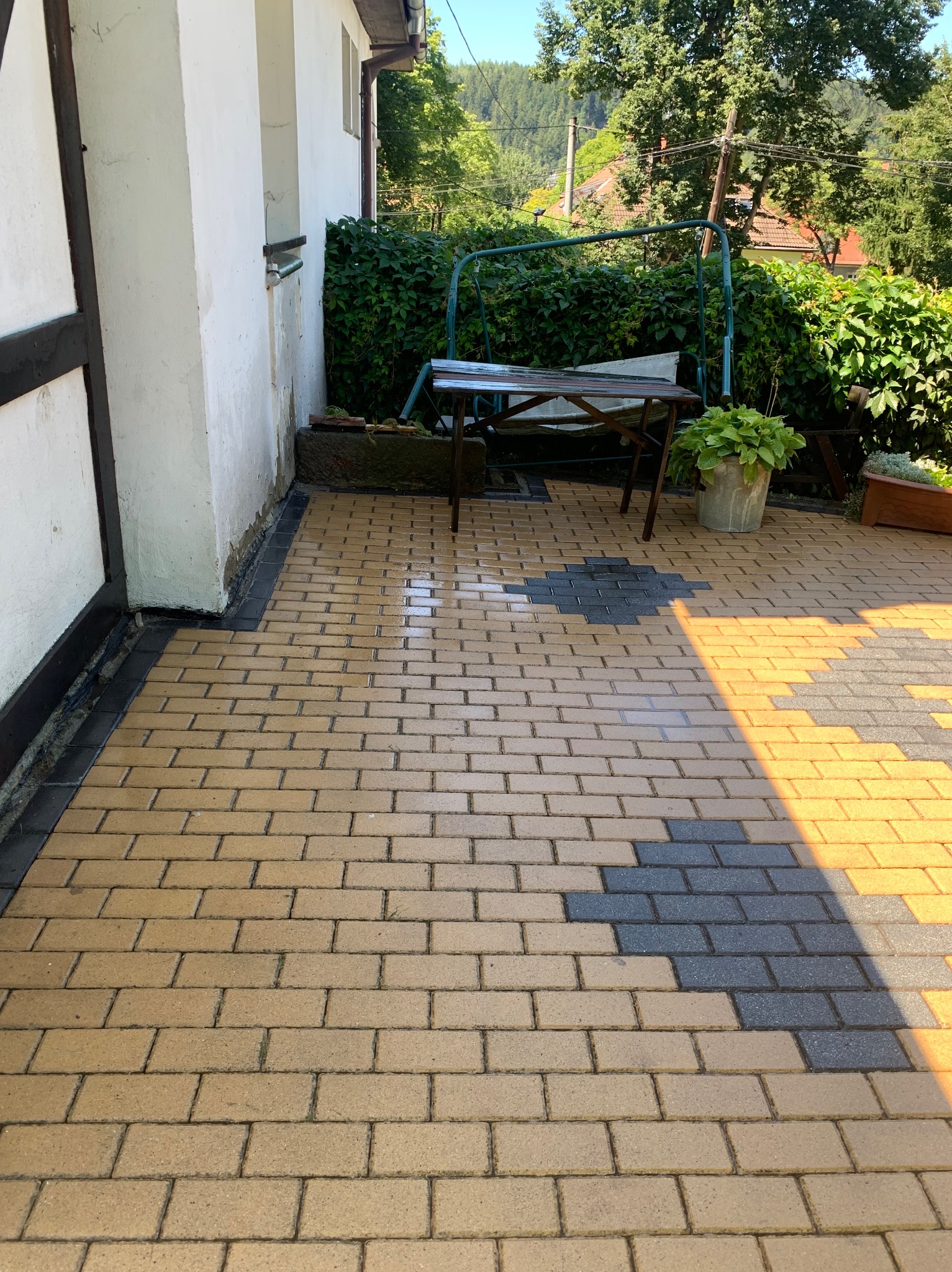 Świeżo umyta, dwukolorowa kostka brukowa przed domem z widocznymi mokrymi plamami i odbiciami światła słonecznego, obok ściany budynku i huśtawki ogrodowej.