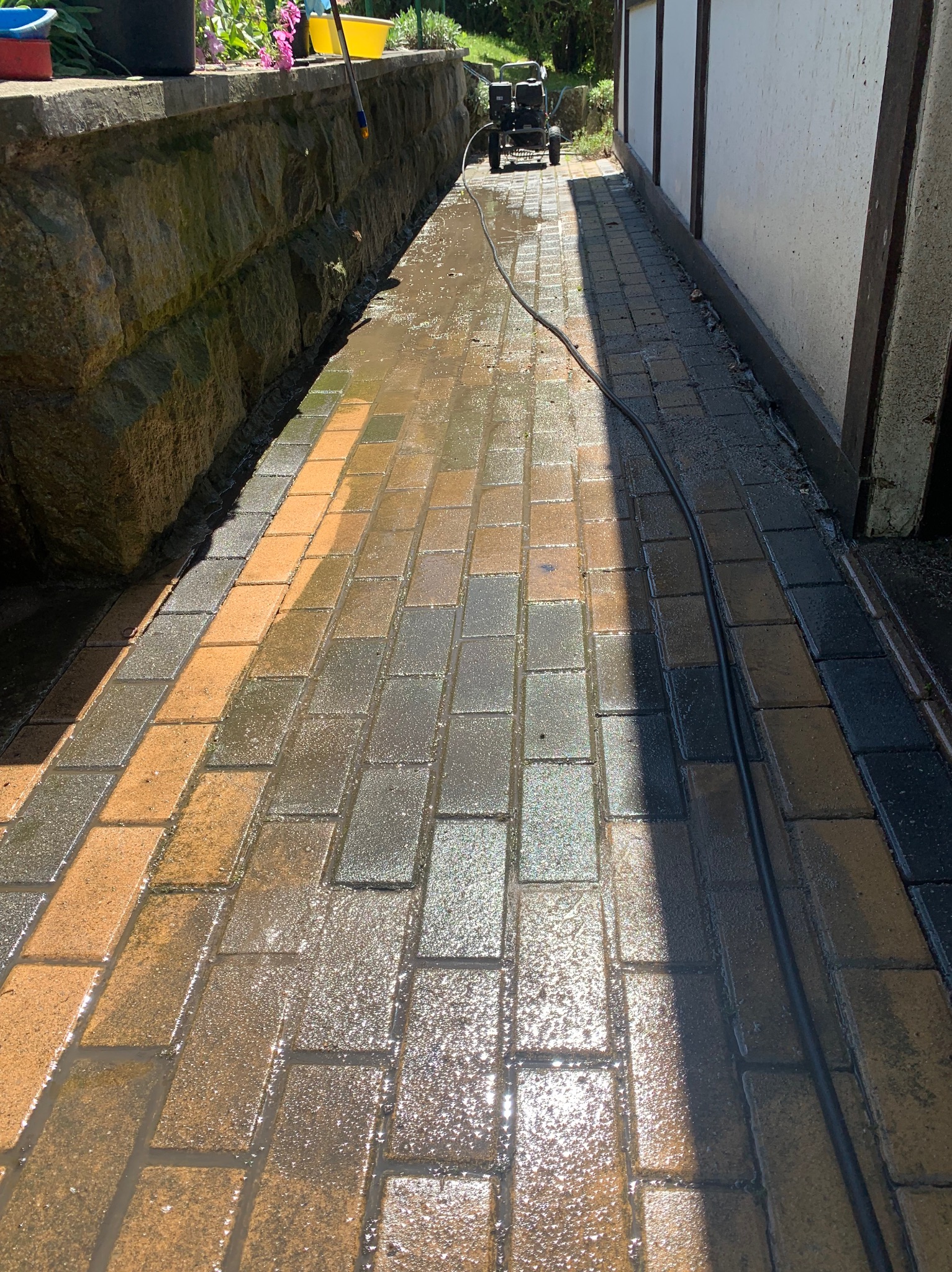 Mokra kostka brukowa w trakcie czyszczenia myjką ciśnieniową, widoczny wąż i urządzenie, część kostki już umyta, kontrast między czystą a brudną powierzchnią.