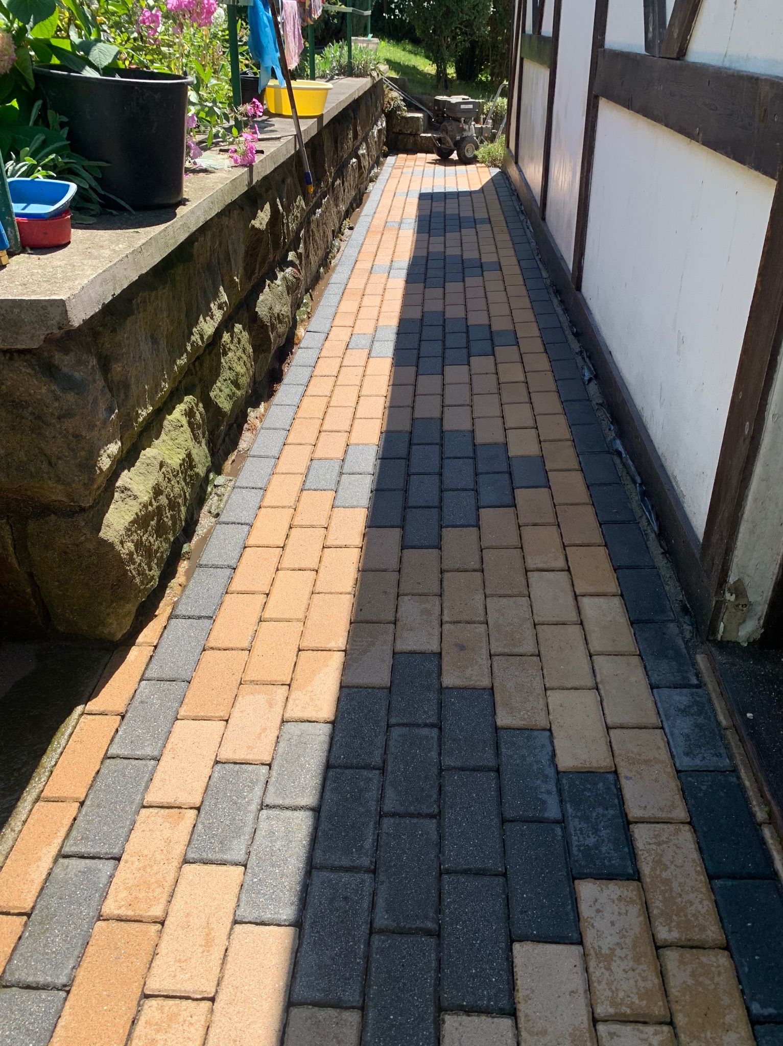 Świeżo umyta, dwukolorowa kostka brukowa w odcieniach szarości i beżu, ułożona wzdłuż ściany budynku z elementami drewnianymi, z widocznymi śladami wilgoci w spoinach i lekkimi cieniami.