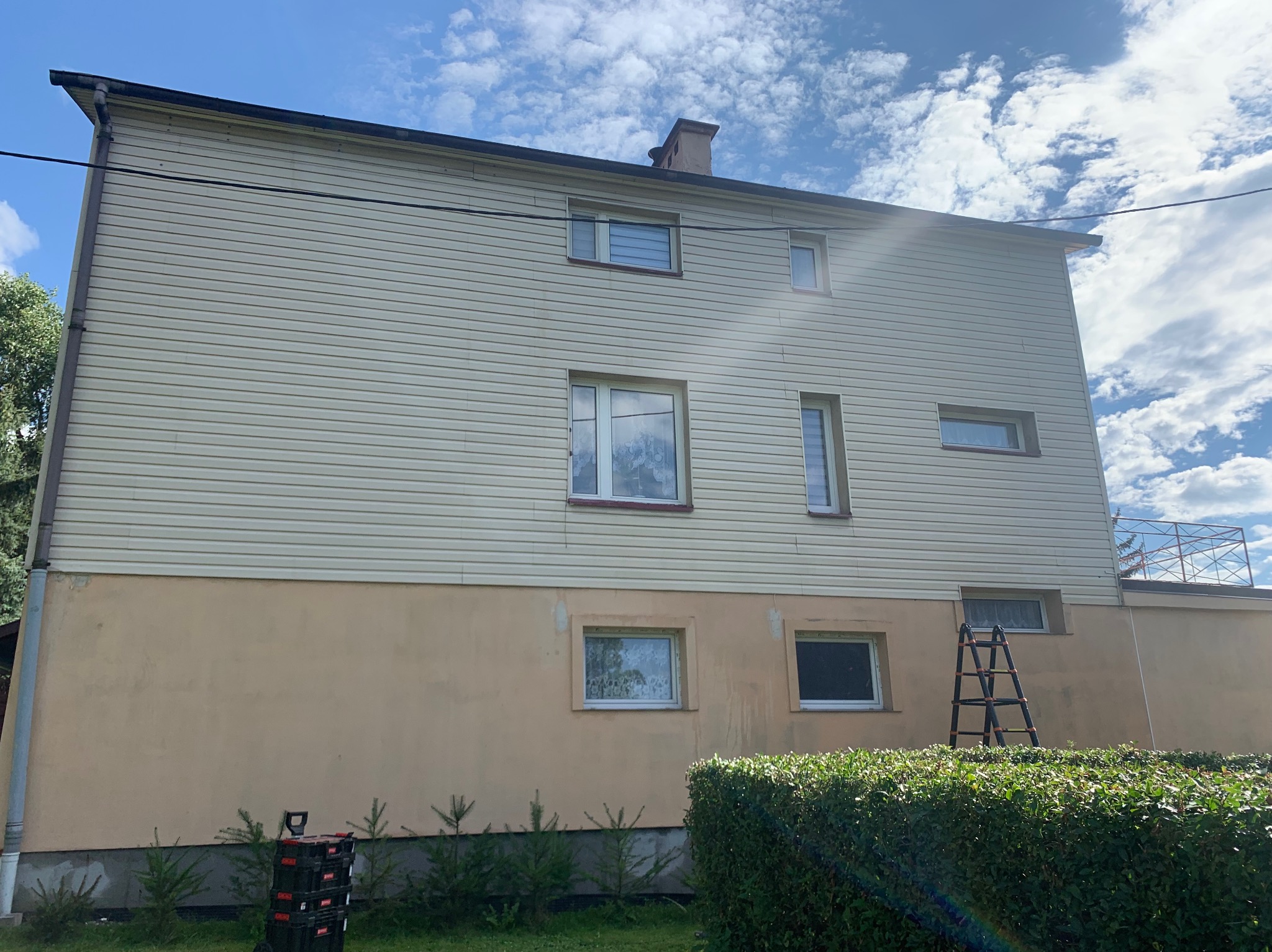 Dwupiętrowy dom z beżową elewacją sidingową i pomalowaną dolną częścią, drabina oparta o ścianę, skrzynki narzędziowe przy podstawie, widok z poziomu ogrodu z krzewami i młodymi drzewkami.