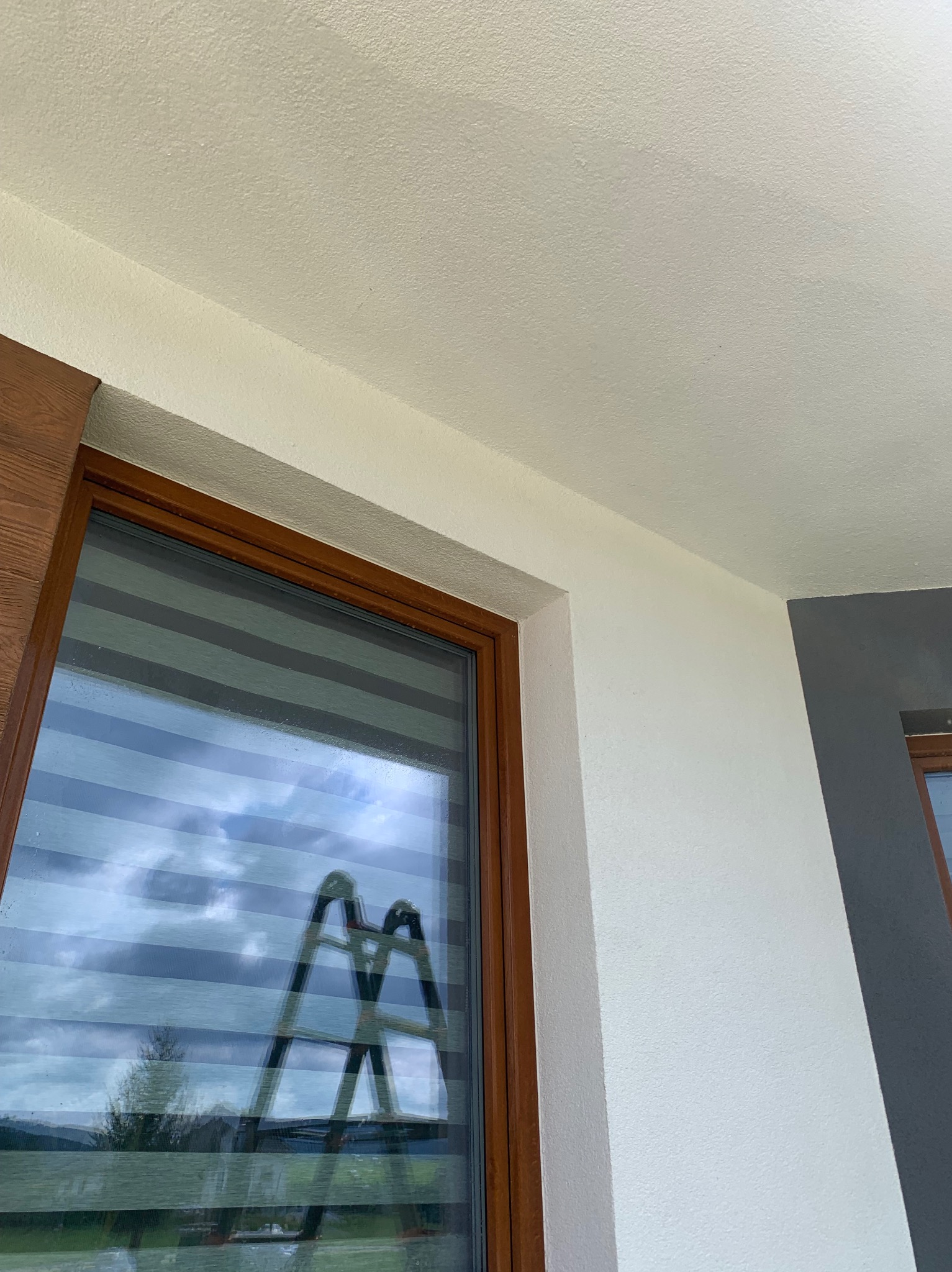 Fragment elewacji budynku z oknem w brązowej ramie, widoczny kawałek drabiny odbijający się w szybie okiennej, ściana pomalowana na biało i szaro.