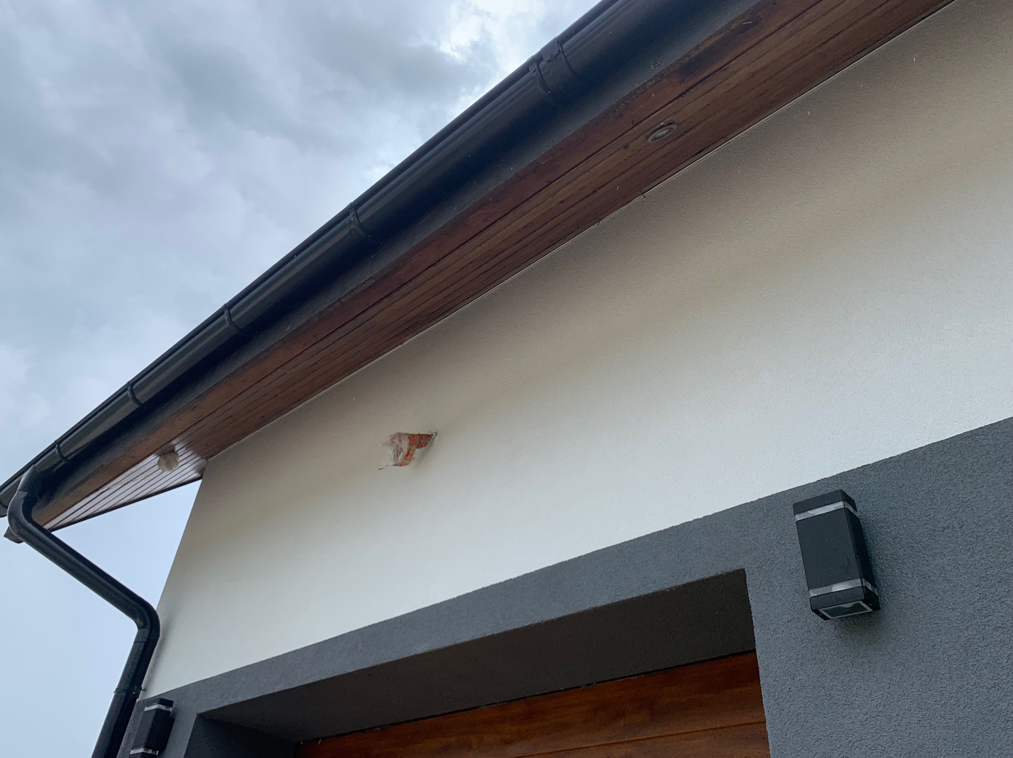 Fragment elewacji budynku z widocznym uszkodzeniem tynku, czarną rynną i nowoczesnym oświetleniem zewnętrznym w pochmurny dzień.