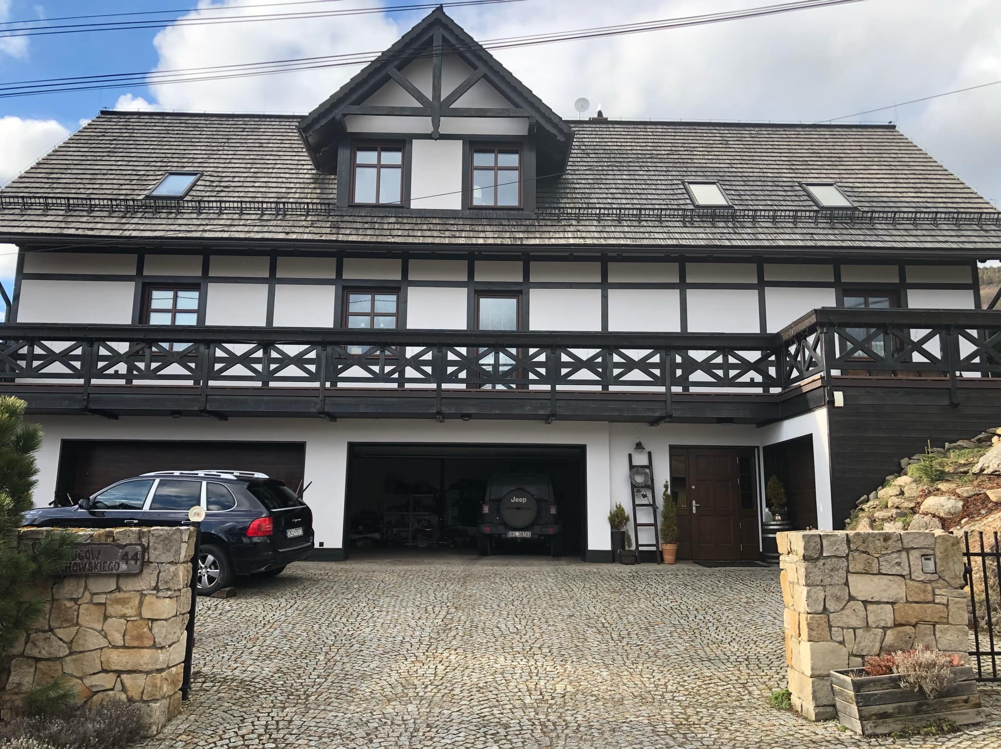 Dwukondygnacyjny dom w stylu szwajcarskim z ciemnym drewnem i białymi ścianami, dach pokryty gontem bitumicznym, podjazd z kostki granitowej, dwa samochody w garażu, numer 44 na kamiennym słupku.