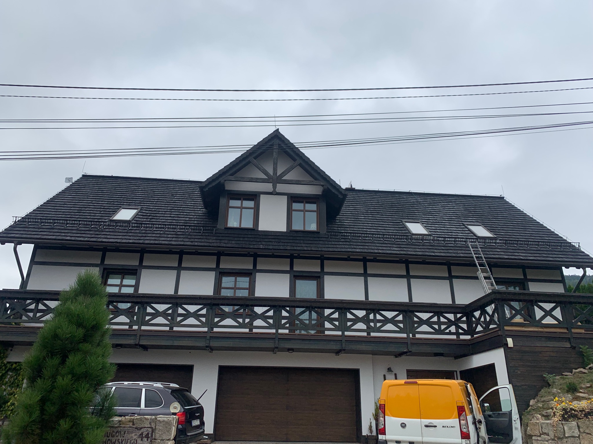 Dwupiętrowy dom z ciemnym dachem, balkonem z drewnianą balustradą i drabiną opartą o dach, zaparkowane dwa samochody, pochmurne niebo.