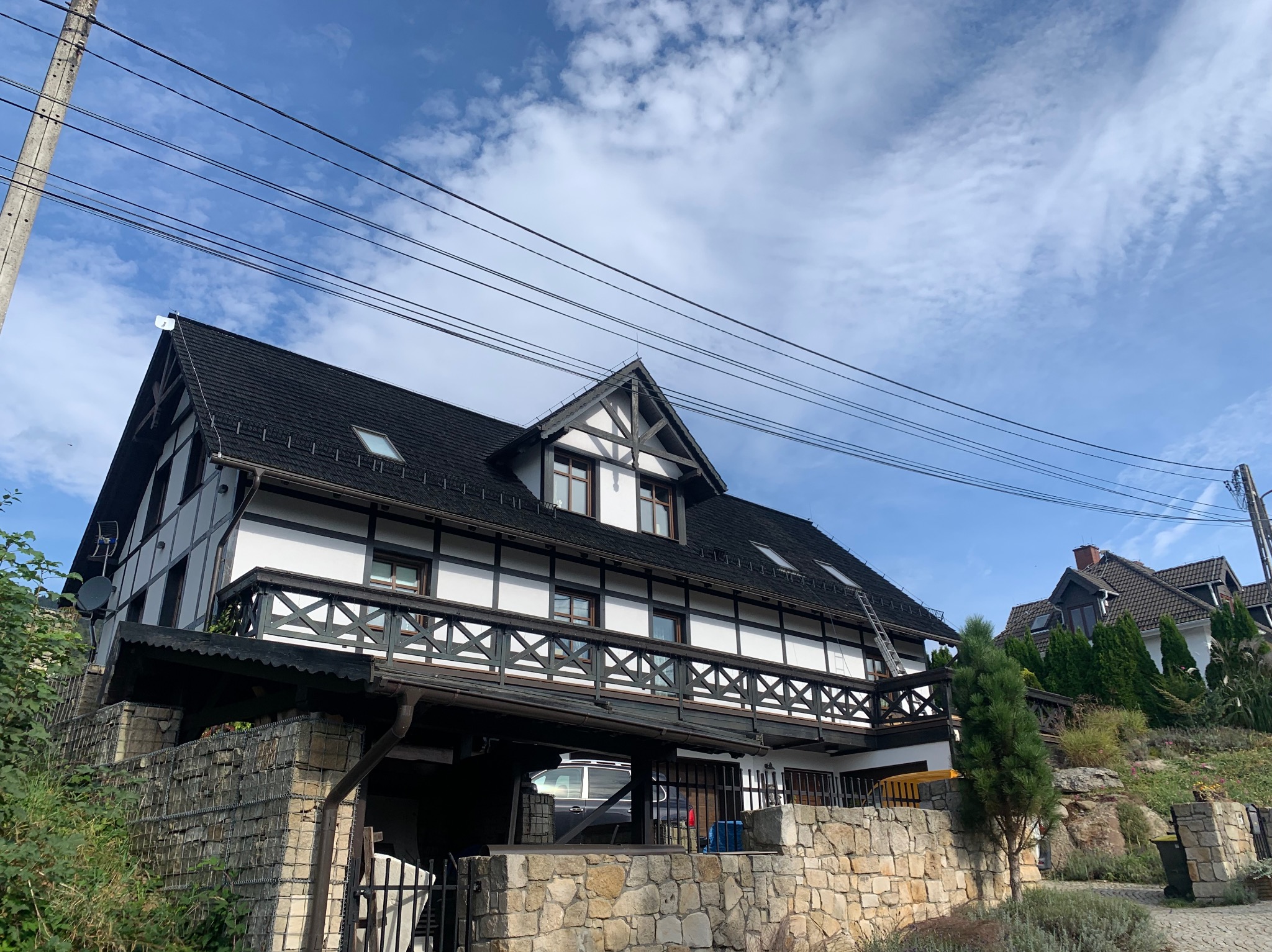 Dwupiętrowy dom w stylu szachulcowym z czarnym dachem i białymi ścianami, widoczny z dołu pod kątem, z drabiny opartą o dach, na tle błękitnego nieba z obłokami.