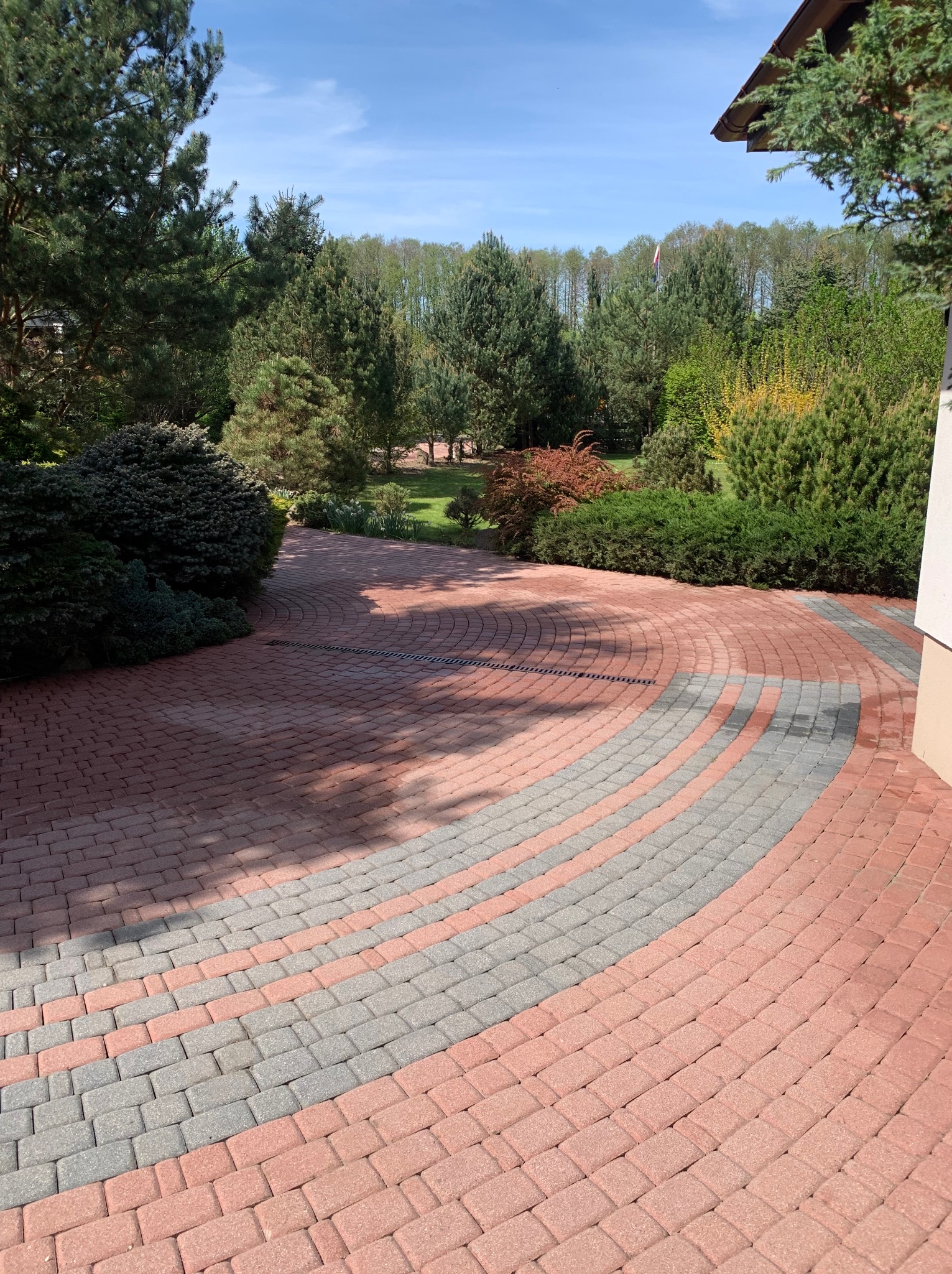 Układanka z czerwono-szarej kostki brukowej tworząca półokrąg na tle zieleni. Widoczny odpływ liniowy i fragment budynku z prawej strony. Słoneczny dzień.