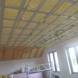 Paweł Stroński - Montaż izolacji z wełny mineralnej na poddaszu, widoczna metalowa konstrukcja pod płyty gipsowo-kartonowe, fragment ściany ze szpachlowaną powierzchnią, drabina rusztowaniowa i okna.