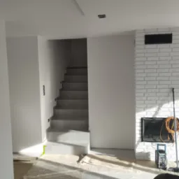 Wnętrze w trakcie remontu: zabezpieczona folią podłoga, białe ściany, schody wyłożone szarą płytką, kominek z białej cegły, widoczne narzędzia i sprzęt budowlany.