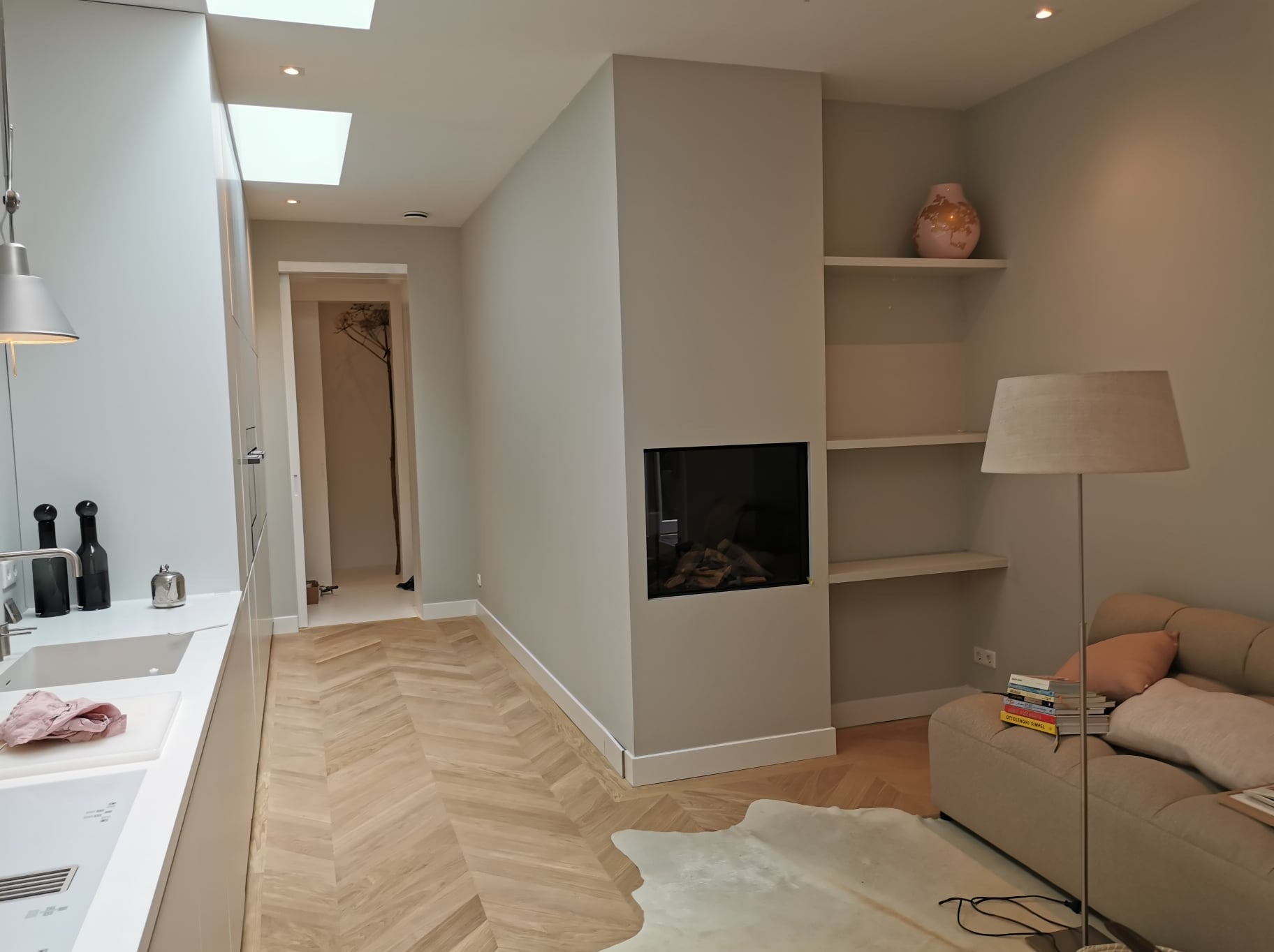 Nowoczesne wnętrze z parkietem ułożonym w jodełkę, minimalistycznym kominkiem, zabudowanymi półkami, sofą i lampą podłogową, doświetlone światłem dziennym z okien dachowych.