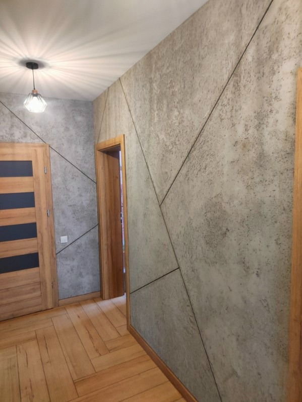 Ściana wykończona dekoracyjną, szarą strukturą z geometrycznym podziałem na panele, drewniane drzwi z czarnymi wstawkami, parkiet ułożony w jodełkę, wisząca lampa z kloszem w stylu retro.
