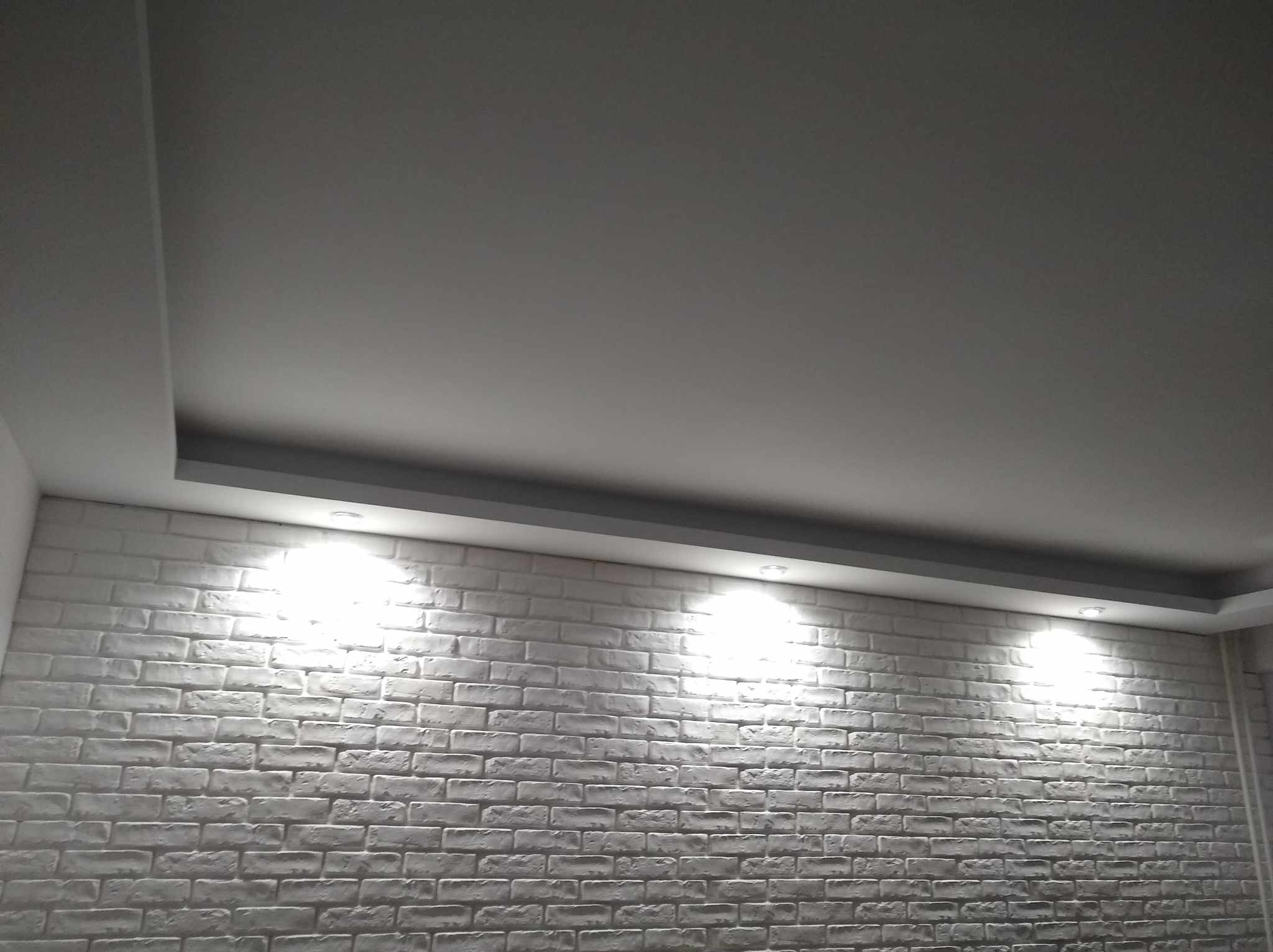 Ściana z białej cegły dekoracyjnej z trzema punktowymi lampami sufitowymi, szara listwa oświetleniowa pod sufitem, widok z dołu.