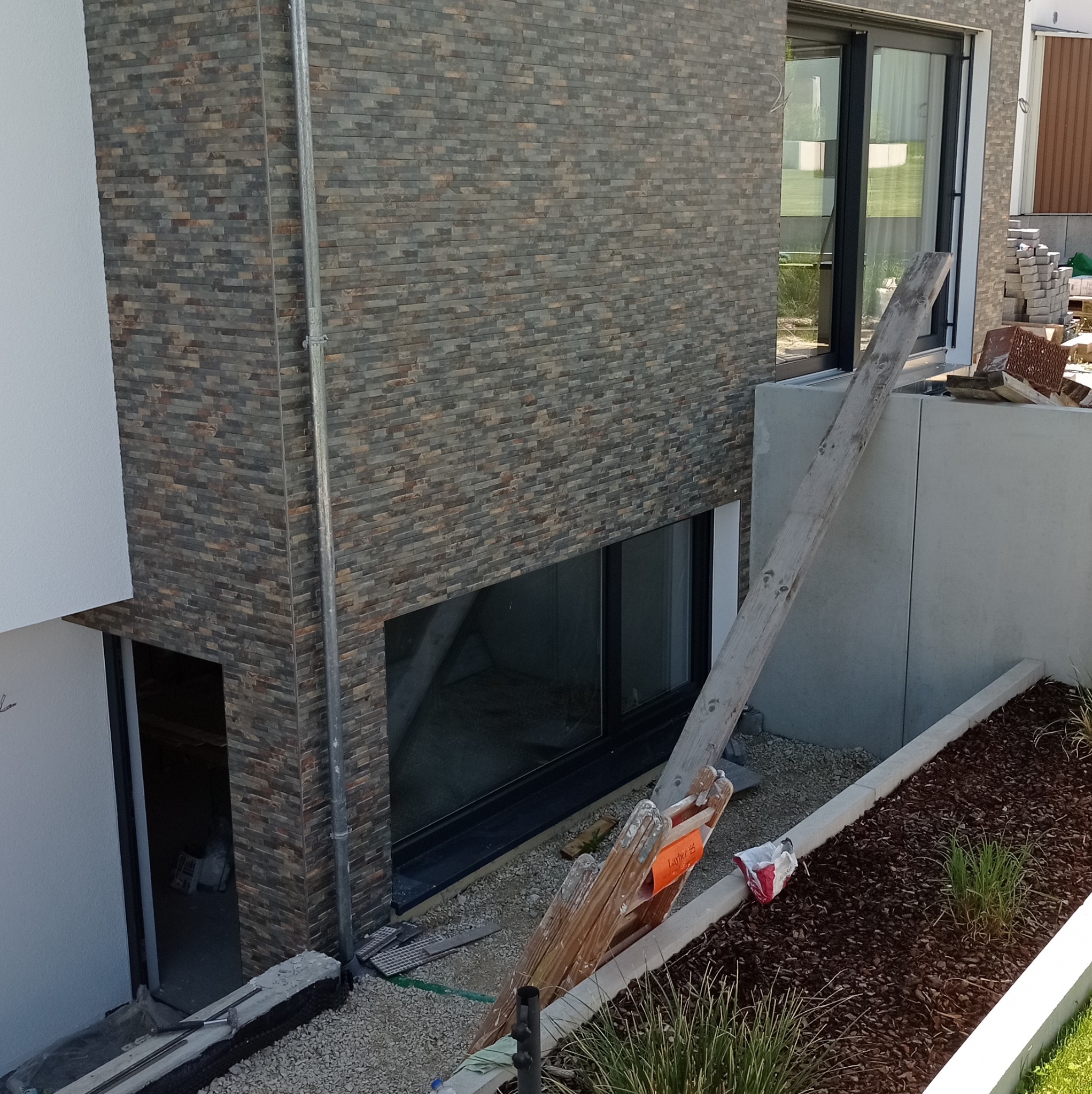 Fragment elewacji budynku wykończonej płytkami imitującymi kamień, z widoczną drabinką opartą o ścianę i fragmentem rabaty z korą i roślinami.