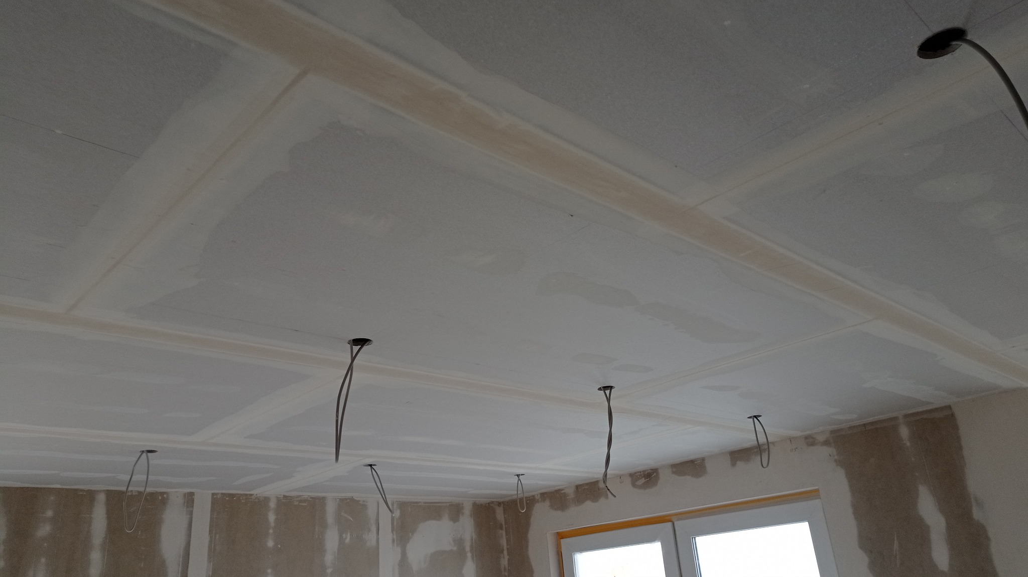 Sufit z płyt kartonowo-gipsowych w trakcie montażu, widoczne łączenia płyt i wystające przewody elektryczne, ściany w surowym stanie przygotowane do dalszych prac wykończeniowych.