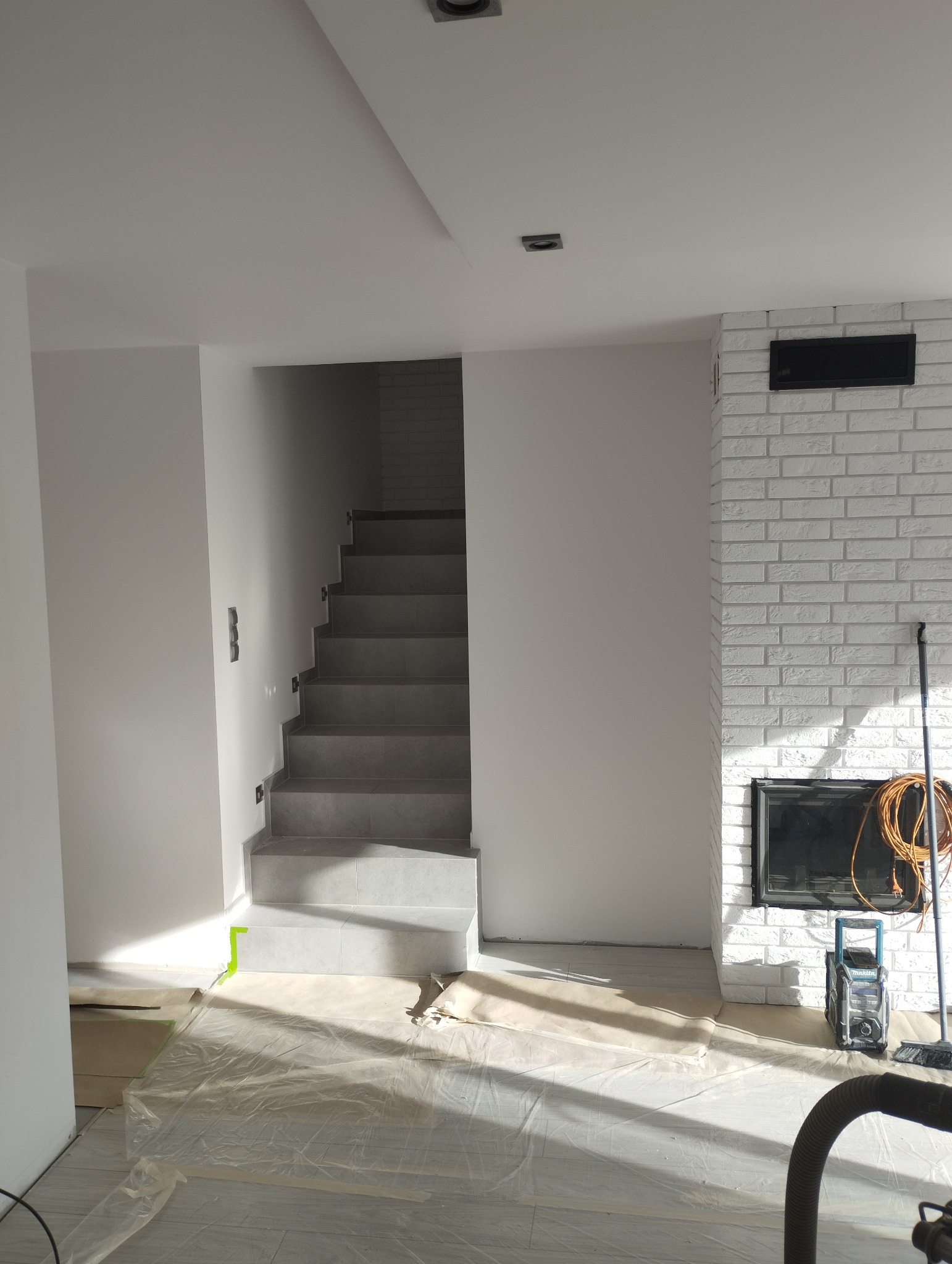 Wnętrze w trakcie remontu: zabezpieczona folią podłoga, białe ściany, schody wyłożone szarą płytką, kominek z białej cegły, widoczne narzędzia i sprzęt budowlany.