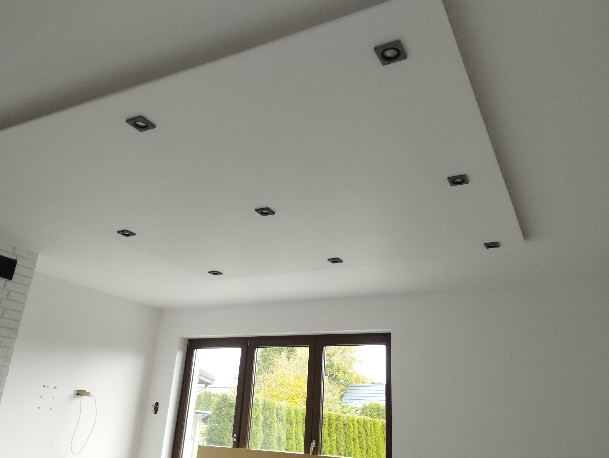 Biały sufit podwieszany z zamontowanymi czarnymi, kwadratowymi oprawami oświetleniowymi, widok na okno z brązową ramą i zielony żywopłot na zewnątrz, ściana z imitacją białej cegły w tle.