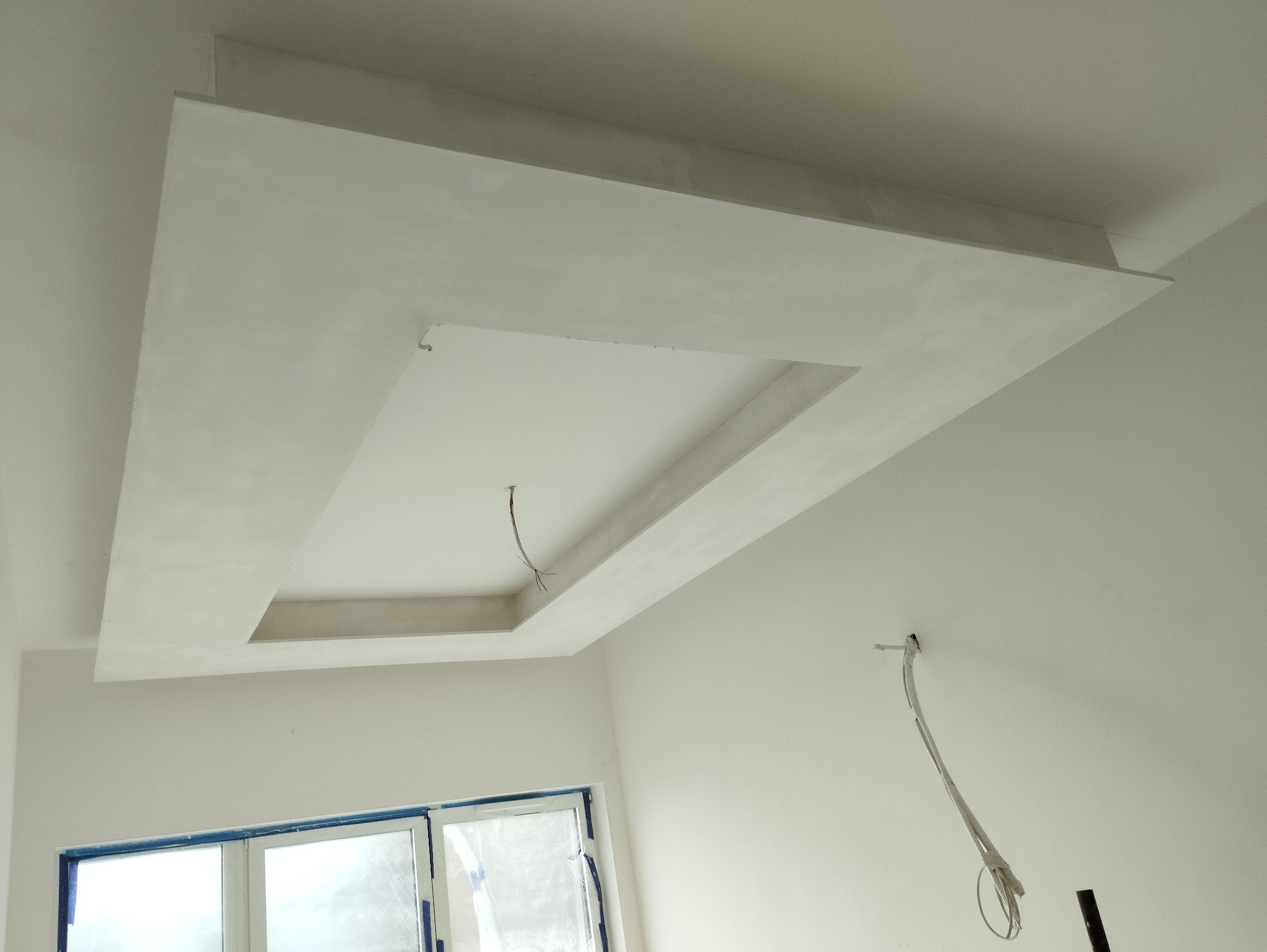 Kaskadowy sufit podwieszany z płyt kartonowo-gipsowych, z centralnym prostokątnym wgłębieniem, w trakcie wykończenia, widoczne przewody elektryczne, okno zabezpieczone folią malarską.