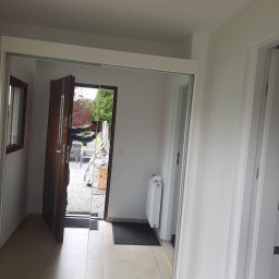 Waldemar Komora - Wnętrze przedpokoju z zamontowaną szafą przesuwną z lustrzanymi drzwiami, odbijającymi otwarte drzwi wejściowe i widok na zewnątrz. Jasne płytki podłogowe.