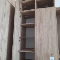 Zamontowana szafa z otwartymi drzwiami i półkami o fakturze drewna. Widoczne zawiasy i etykiety na wewnętrznych ścianach. Ujęcie z bliska, w neutralnym oświetleniu.