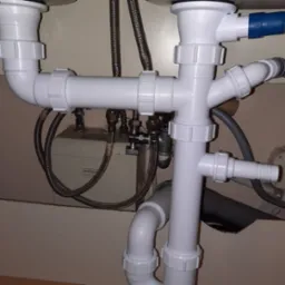 Złożony system białych rur odpływowych pod zlewem, z widocznymi elementami mocującymi i metalowymi wężykami, zainstalowany w szafce z kremowym tłem.