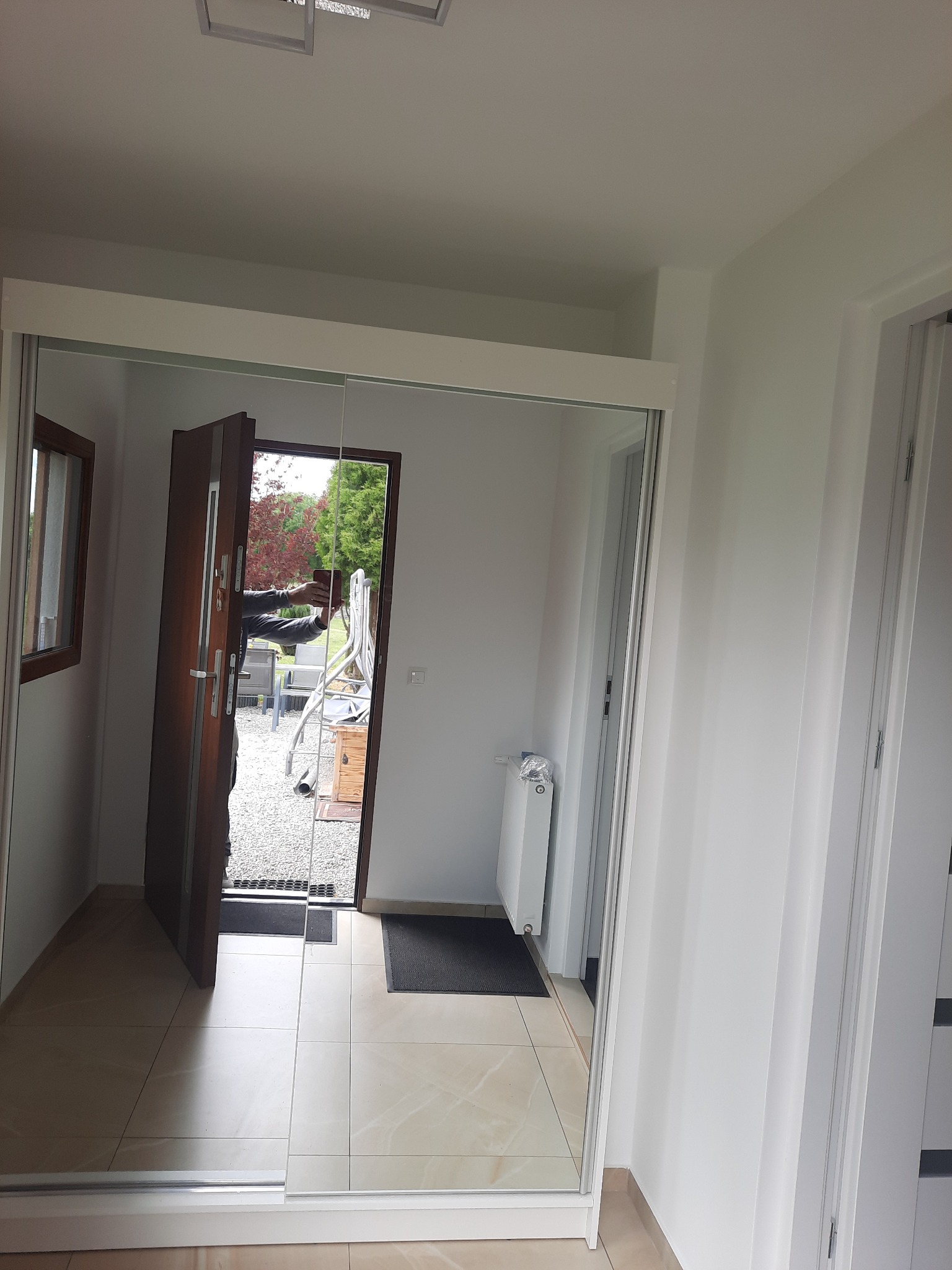 Wnętrze przedpokoju z zamontowaną szafą przesuwną z lustrzanymi drzwiami, odbijającymi otwarte drzwi wejściowe i widok na zewnątrz. Jasne płytki podłogowe.